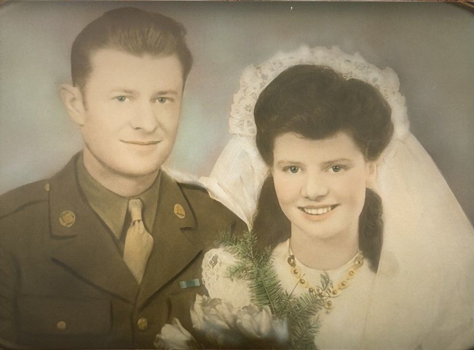A man in a military uniform poses next to a woman in a wedding dress and veil, both smiling at the camera. The woman holds a bouquet of flowers.