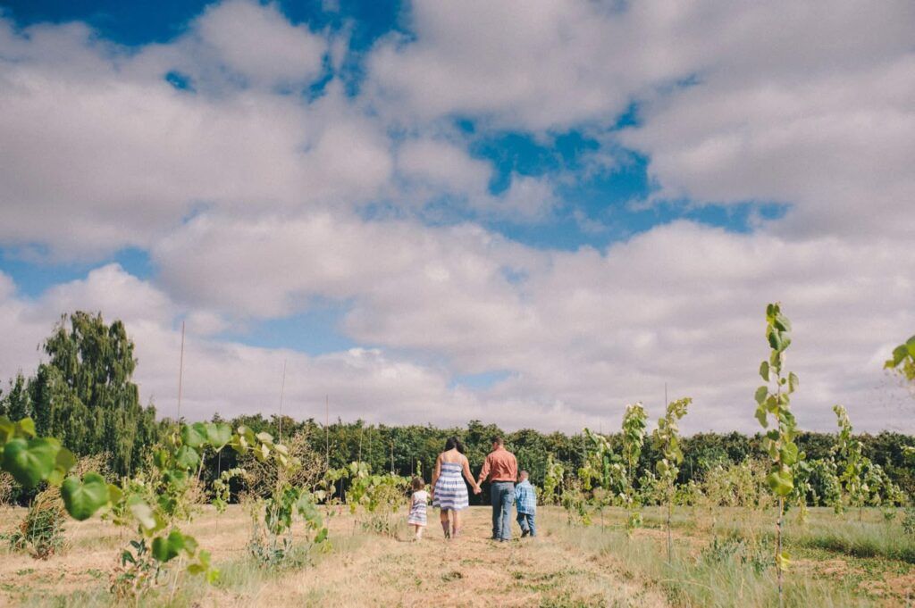 Three people, two adults and a child, walk hand in hand through a field with young plants under a partly cloudy sky.
