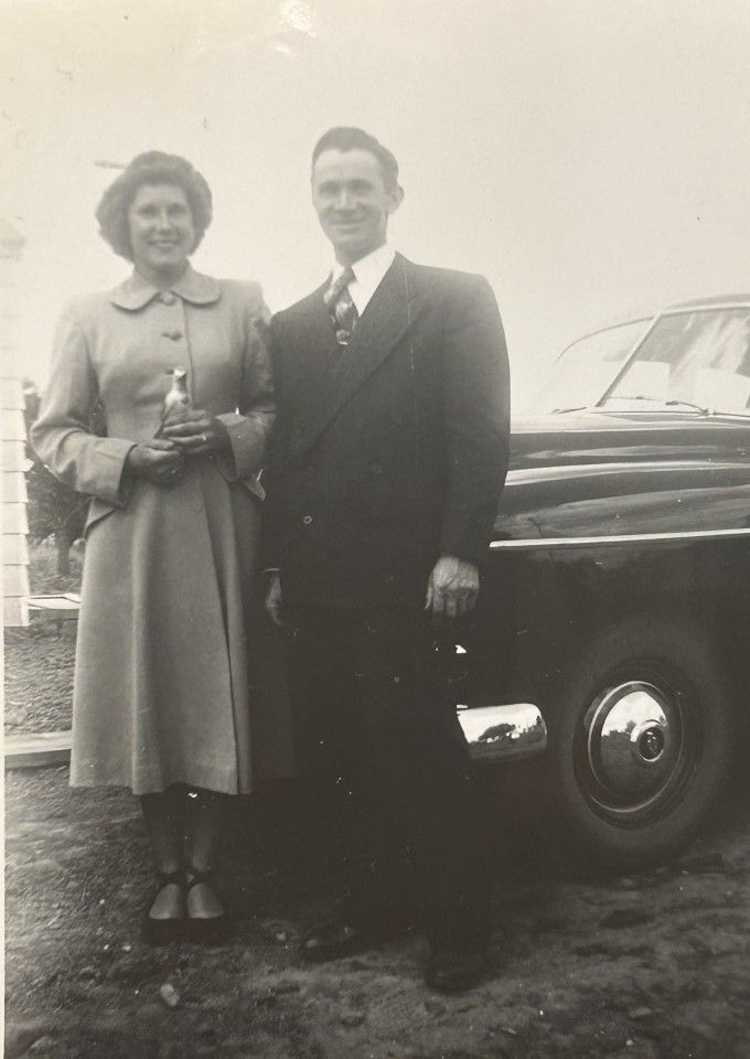 A woman in a long coat and a man in a suit stand next to an old car, posing for a black and white photograph.